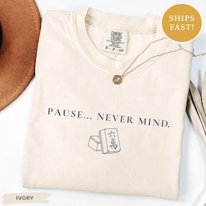 May include: An ivory short-sleeve t-shirt featuring the dark blue text PAUSE... NEVER MIND. and a graphic illustration of two mahjong tiles. The shirt has a crew neckline and a Comfort Colors brand label printed on the inside of the collar.