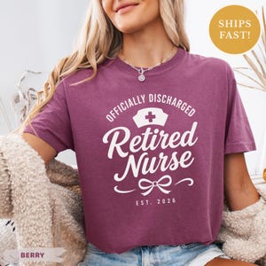 May include: A berry-colored short-sleeve t-shirt featuring white screen-printed text that reads "OFFICIALLY DISCHARGED" above a nurse cap icon, the words "Retired Nurse" in a script font, and "EST. 2026" at the bottom with a small bow graphic.