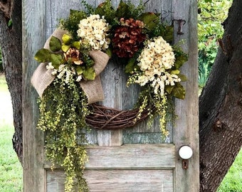 May include: A decorative wreath with a grapevine base, adorned with cream and burgundy hydrangeas, green leaves, and trailing greenery. A burlap bow adds texture. The wreath hangs on a weathered, light blue wooden door.