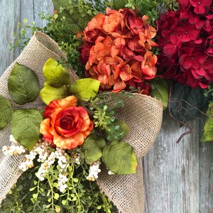 May include: A decorative wreath featuring a large burlap ribbon bow, deep red and burnt orange hydrangea blooms, a single orange rose, green foliage, and clusters of small white berries. The arrangement is set against a weathered wooden surface.