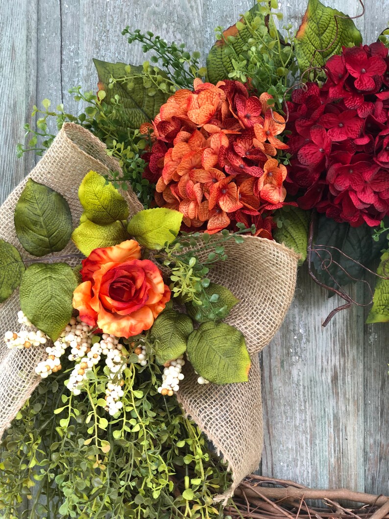 May include: A decorative wreath featuring a large burlap ribbon bow, deep red and burnt orange hydrangea blooms, a single orange rose, green foliage, and clusters of small white berries. The arrangement is set against a weathered wooden surface.