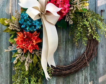 May include: A wreath made of grapevine with a large bow of cream-colored ribbon. The wreath is decorated with artificial flowers in shades of pink, orange, and blue. The wreath is hanging on a weathered wooden door.