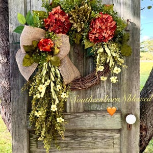 May include: A wreath with a burlap bow and a cascade of white flowers. The wreath is decorated with red and green hydrangeas and a green flower. The wreath is hanging on a wooden door with the text "Southernborn Blessed" written on it.