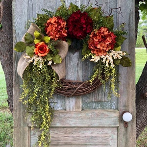 May include: A decorative wreath featuring deep red and burnt orange hydrangea blooms, a single orange rose, and green foliage. The arrangement includes a natural burlap ribbon bow and trailing vines attached to a circular brown woven twig base.