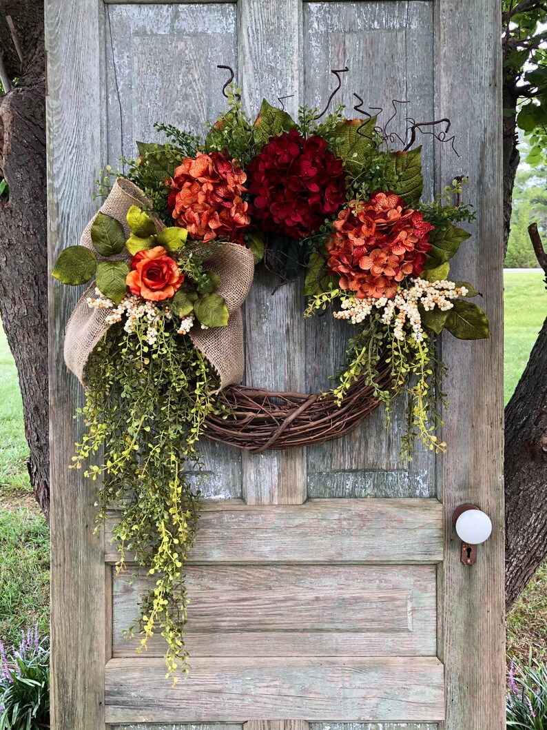 May include: A decorative wreath featuring deep red and burnt orange hydrangea blooms, a single orange rose, and green foliage. The arrangement includes a natural burlap ribbon bow and trailing vines attached to a circular brown woven twig base.
