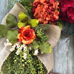 May include: A decorative wreath featuring a large burlap bow, orange and deep red hydrangea blooms, a single orange rose, green foliage, and clusters of small white berries. The arrangement is set against a base of natural brown twigs.