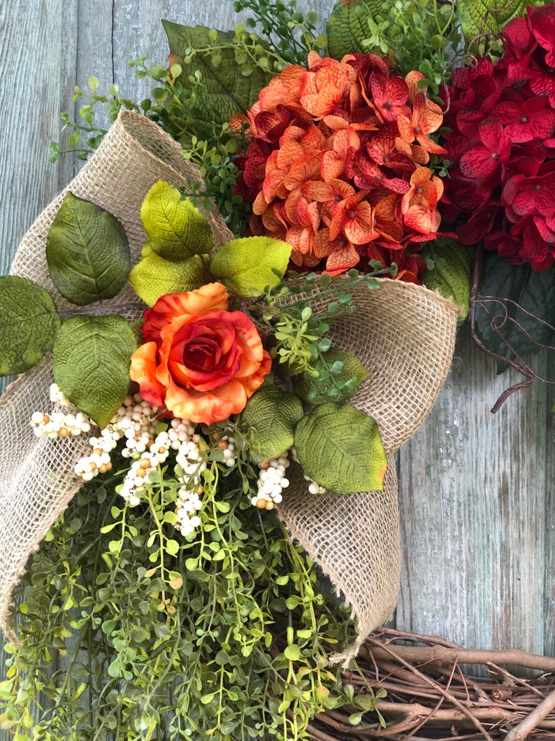 May include: A decorative wreath featuring a large burlap bow, orange and deep red hydrangea blooms, a single orange rose, green foliage, and clusters of small white berries. The arrangement is set against a base of natural brown twigs.