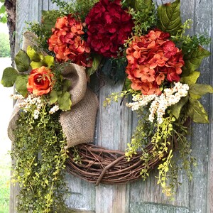 May include: A decorative wreath featuring a natural grapevine base adorned with deep red and burnt orange hydrangea blooms, a single orange rose, green foliage, white berry clusters, and a wide tan burlap ribbon bow with long trailing greenery.