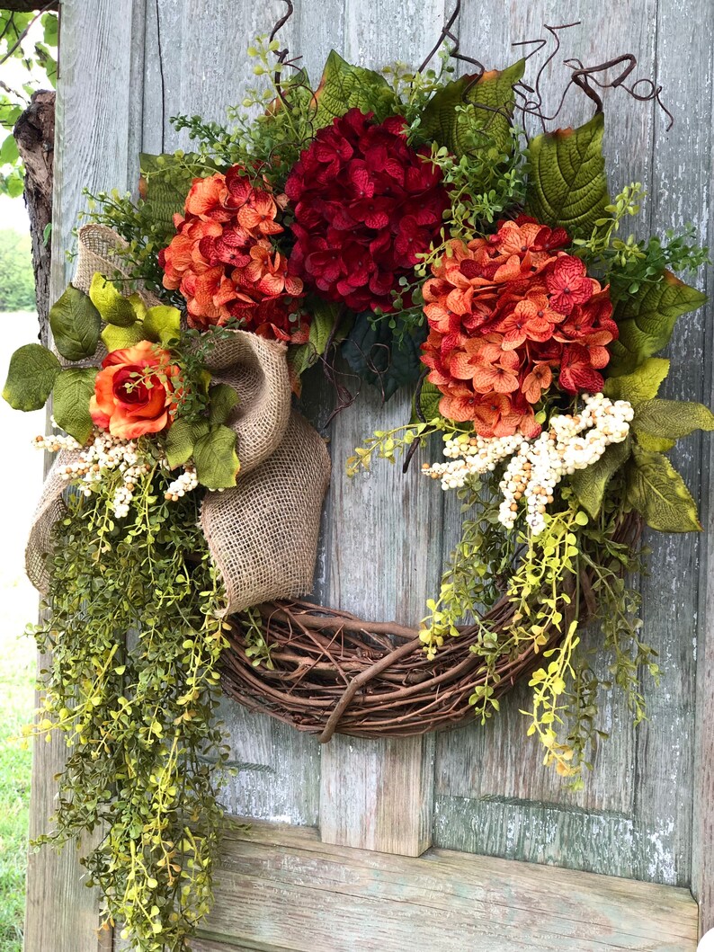 May include: A decorative wreath featuring a natural grapevine base adorned with deep red and burnt orange hydrangea blooms, a single orange rose, green foliage, white berry clusters, and a wide tan burlap ribbon bow with long trailing greenery.