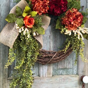 May include: A decorative wreath featuring a natural grapevine base adorned with artificial orange and deep red hydrangea blooms, a single orange rose, green foliage, white berry clusters, and a large tan burlap bow with long trailing greenery.