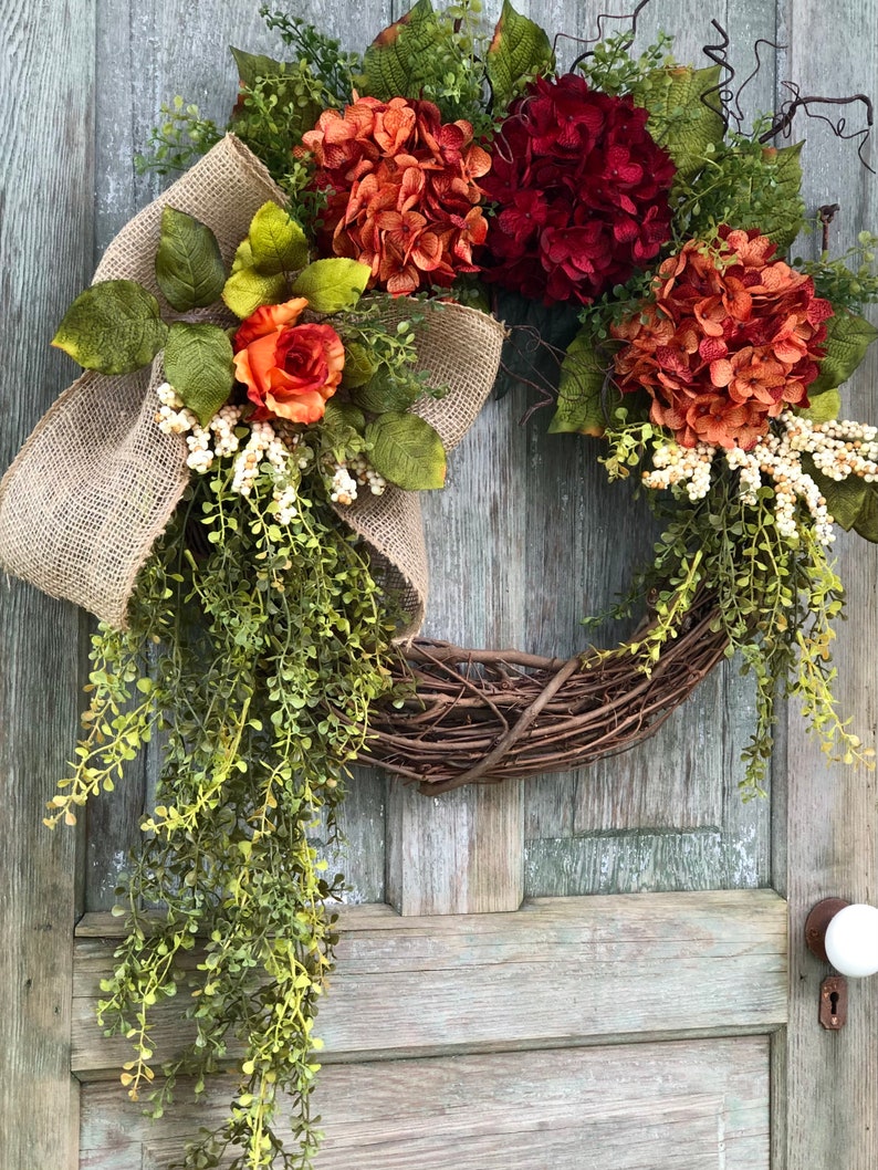 May include: A decorative wreath featuring a natural grapevine base adorned with artificial orange and deep red hydrangea blooms, a single orange rose, green foliage, white berry clusters, and a large tan burlap bow with long trailing greenery.