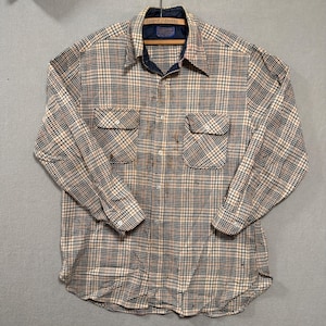 May include: A long-sleeved plaid shirt in beige, navy, and rust. The shirt has two chest pockets and a button-down front. The collar and inner cuffs are navy. The shirt is hanging on a wooden hanger.