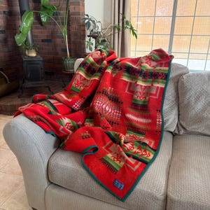 May include: A red wool blanket featuring a geometric pattern in shades of green, black, and cream. The blanket has a dark green border and a small rectangular label in the corner. The design includes repeating angular shapes and lines.