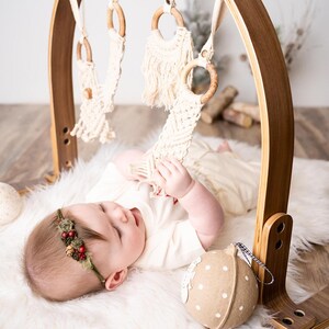 May include: A wooden baby gym with hanging macrame toys and wooden rings. A baby wearing a floral headband and a white onesie is lying on a white fluffy rug, reaching for the toys. A decorative felt ball is also present.
