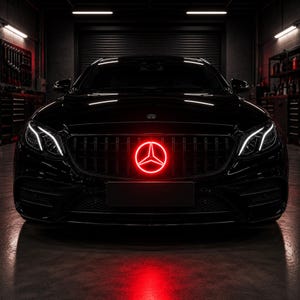 May include: A black Mercedes-Benz sedan viewed from the front, featuring a vertical slat grille and a prominent, glowing red circular three-pointed star emblem centered on the front grille, with bright white LED daytime running lights illuminated.