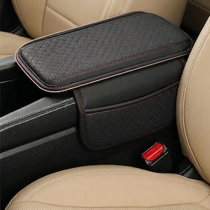 May include: Black car armrest cover with a diamond pattern and red stitching. The cover is designed to fit over the center console armrest. The interior of the car has tan leather seats.