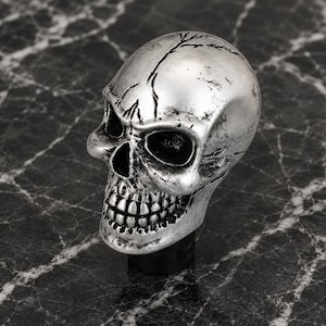 May include: A silver-coloured gear shift knob shaped like a human skull. The skull features a metallic finish with dark, weathered detailing in the recessed areas of the eye sockets, nasal cavity, and teeth, mounted on a black cylindrical base.