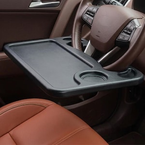 May include: A black car steering wheel tray with a rectangular surface and two cup holders. The tray is designed to attach to a car's steering wheel for use as a desk or table. The car's interior features brown leather seats and a brown steering wheel.