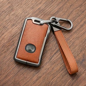 Peut inclure: Un étui de porte-clés en cuir marron avec un cadre en métal et une dragonne en cuir assortie. L'étui a un fermoir en métal gris foncé et un petit emblème gris foncé. L'étui est conçu pour protéger et transporter les clés.