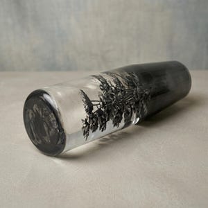May include: A clear glass vase with a black, tree-like design on one side. The vase transitions from clear to a dark, smoky gray at the top. The vase is cylindrical with a rounded base.