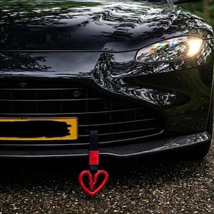 May include: A black luxury car with a red heart-shaped tow hook. The car's front features a black grille, headlights, and a license plate. The tow hook is attached to the car's front bumper.