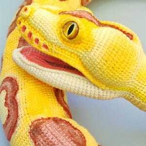 May include: A yellow snake plush toy with brown markings. The snake's head is detailed with a red mouth and yellow eyes. The toy is made of a textured material, and is likely handmade.