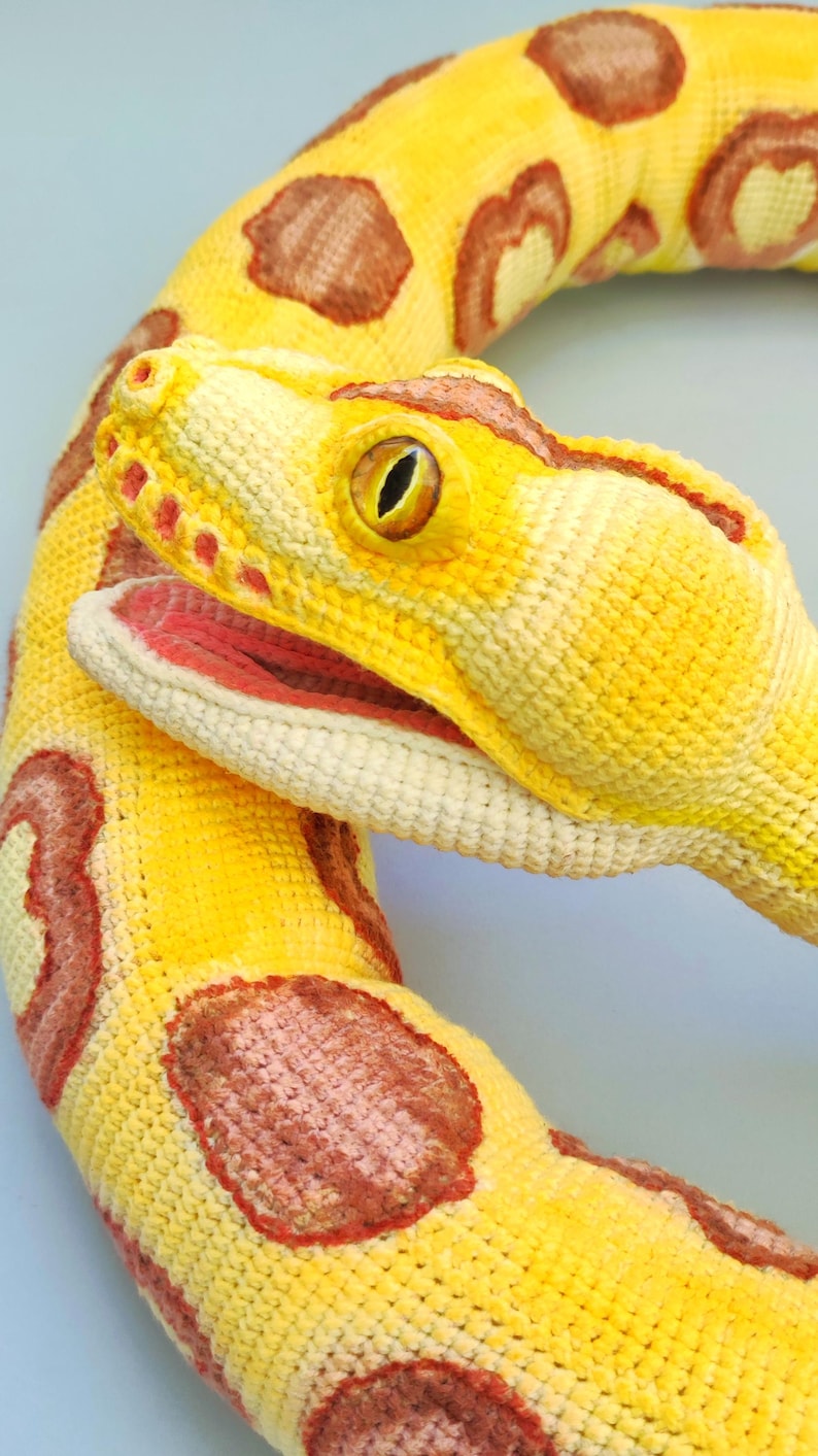 May include: A yellow snake plush toy with brown markings. The snake's head is detailed with a red mouth and yellow eyes. The toy is made of a textured material, and is likely handmade.