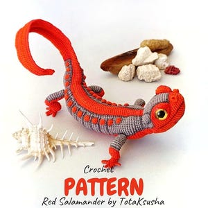 May include: A crocheted red and gray salamander toy with a long curved tail, textured back, and glossy eyes. The text below reads: Crochet PATTERN Red Salamander by TotaKsusha. The salamander is positioned near a white seashell and small rocks.