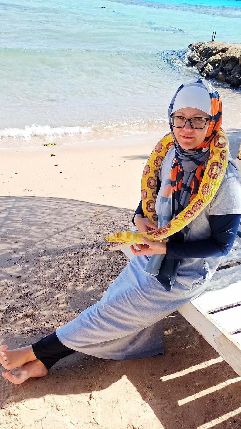 May include: A person wearing glasses and a headscarf is holding a yellow and brown toy snake. The snake is wrapped around the person's neck. The person is sitting on a white bench near the beach.