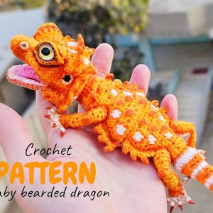 May include: A crocheted baby bearded dragon in shades of orange and white, with a pink mouth and detailed features. The text "Crochet PATTERN Baby bearded dragon" is displayed on the image. The dragon is held in a hand.
