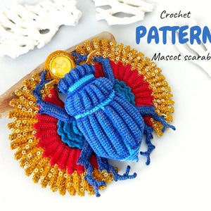 Może przedstawiać: Niebieski szydełkowy skarabeusz z czerwoną i złotą podstawą w kształcie wachlarza. Tekst "Crochet Pattern Mascot scarab" jest wyświetlany na niebiesko. Żuk umieszczony jest na kawałku drewna wyrzuconego na brzeg.
