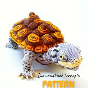 May include: A crocheted diamondback terrapin turtle with a brown and orange shell, gray legs with black spots, and a gray head. The turtle has yellow eyes and claws. The text "Diamondback terrapin PATTERN by TotakSusha" is displayed.
