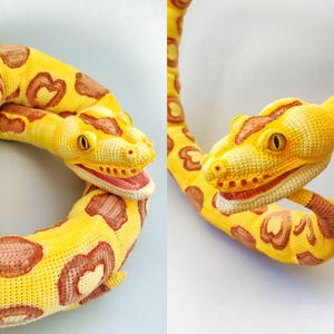 May include: A crocheted snake in yellow and brown. The snake has a detailed head with yellow eyes and a red mouth. The body is yellow with brown heart-shaped markings. The snake is coiled in a circular shape.