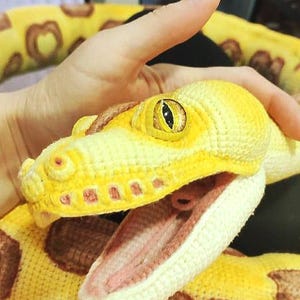 May include: A crocheted snake in shades of yellow and brown, with an open mouth revealing a pink interior. The snake's eye is yellow with a black pupil. The body features a pattern of brown spots. The snake is handmade.