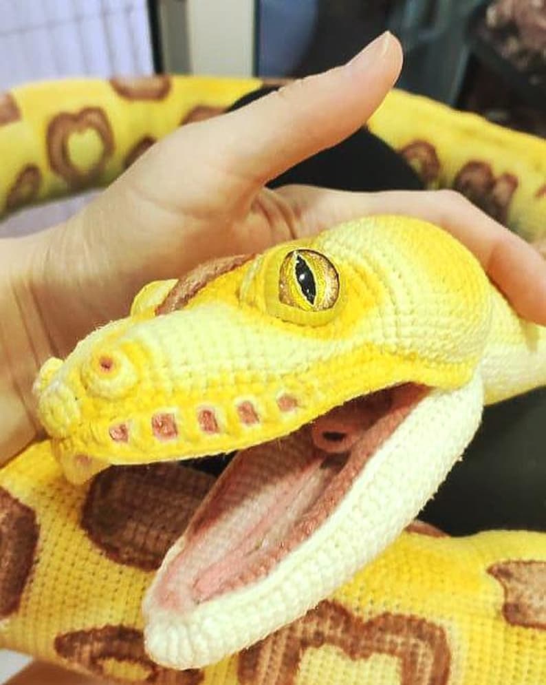 May include: A crocheted snake in shades of yellow and brown, with an open mouth revealing a pink interior. The snake's eye is yellow with a black pupil. The body features a pattern of brown spots. The snake is handmade.