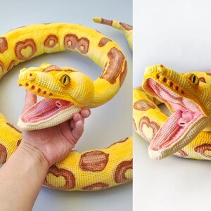 May include: A crocheted yellow snake toy with brown markings and an open mouth showing a pink interior. The snake has yellow eyes and is designed to look realistic. The toy is made of soft yarn.