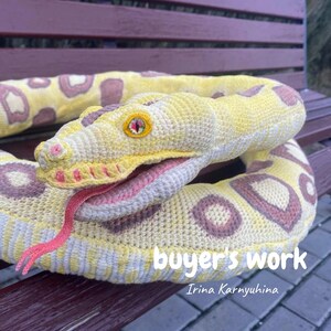May include: A crocheted snake toy in yellow, white, and purple. The snake has a pink tongue and a detailed eye. The text "buyer's work" and the name "Irina Karnyuhina" are visible on the image.