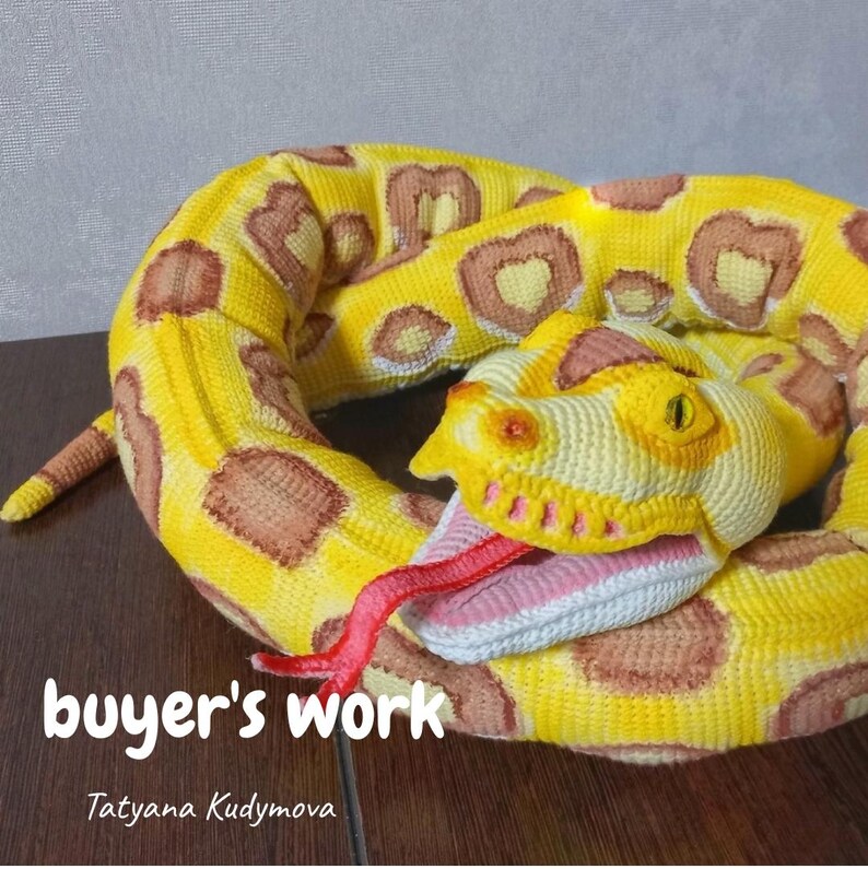May include: A crocheted snake toy in yellow with brown markings. The snake has a detailed head with a pink tongue and open mouth. The text "buyer's work" and the name "Tatyana Kudymova" are visible. A soft toy.
