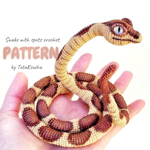 Puede incluir: Una serpiente de ganchillo con manchas marrones y cuerpo beige. La cabeza de la serpiente está levantada y tiene ojos azules. El texto "Snake with spots crochet PATTERN by TotaKsusha" es visible.