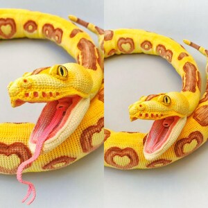May include: A crocheted snake in yellow and brown, featuring heart-shaped patterns. The snake's mouth is open, showing a pink interior and a pink tongue. The eyes are yellow.