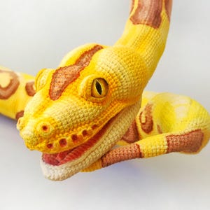 May include: A handcrafted crochet snake in yellow, brown, and cream. The snake's head is detailed with an open mouth, visible red interior, and a yellow eye. The body features brown and yellow patterns.
