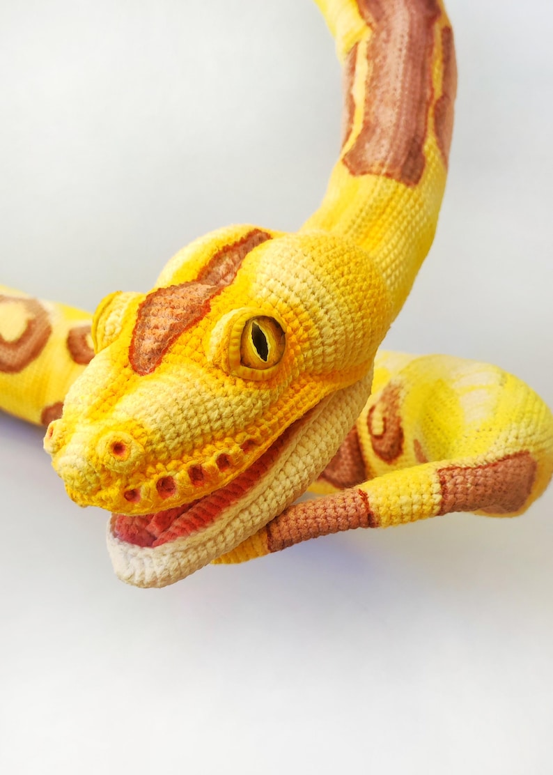 May include: A handcrafted crochet snake in yellow, brown, and cream. The snake's head is detailed with an open mouth, visible red interior, and a yellow eye. The body features brown and yellow patterns.