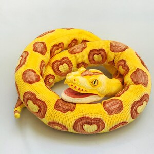 May include: A coiled, plush snake toy in shades of yellow and brown. The snake's body is primarily yellow with brown, heart-shaped markings. The head features an open mouth with a pink interior and a yellow eye. The toy is designed to resemble a snake.