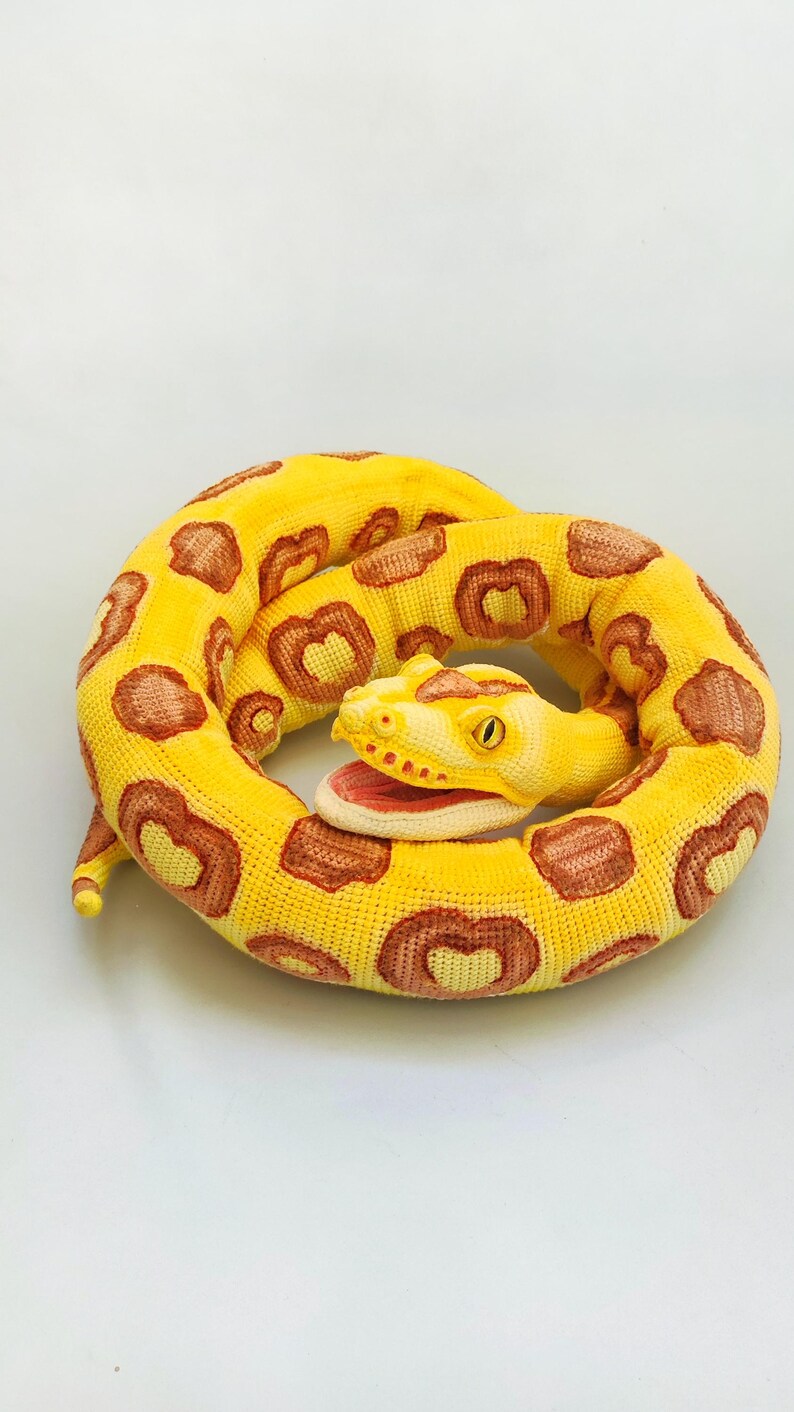May include: A coiled, plush snake toy in shades of yellow and brown. The snake's body is primarily yellow with brown, heart-shaped markings. The head features an open mouth with a pink interior and a yellow eye. The toy is designed to resemble a snake.