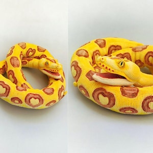 May include: A coiled plush snake in shades of yellow and brown. The snake's body is a vibrant yellow with brown heart-shaped markings. The snake's head is detailed with yellow eyes and a red mouth.