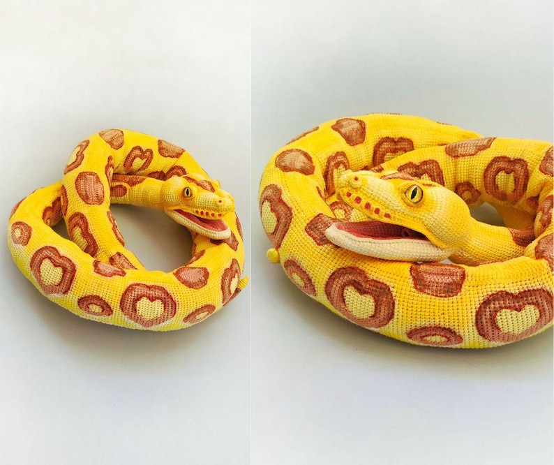 May include: A coiled plush snake in shades of yellow and brown. The snake's body is a vibrant yellow with brown heart-shaped markings. The snake's head is detailed with yellow eyes and a red mouth.
