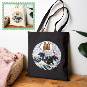 May include: Black tote bag featuring a circular design with a Godzilla-like creature in a wave, a thought bubble with two monkeys, and red hearts. The design is reminiscent of Japanese art.