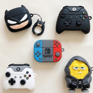 May include: Collection of protective cases for wireless earbuds. Designs include a black Batman, a black Xbox controller, a Nintendo Switch, a white and gray Xbox controller, and a SpongeBob SquarePants character in a gray hoodie.