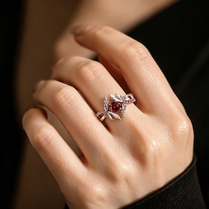 May include: A silver-toned ring featuring a central round deep red stone set within a metal frame shaped like leaves. The band includes small pink accent stones along one side, with the metal leaves extending outward from the center stone.