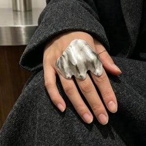 May include: A large, silver-toned ring with an organic, flowing design. The ring has a textured surface and a unique, abstract shape, resembling a cloud or wave. The ring is worn on a hand, with a dark gray coat in the background.